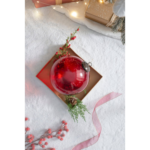 Decorative red glass ornament with greenery on a white surface with Christmas decorations.