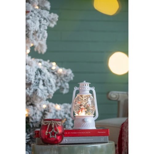 Decorative lantern with a Christmas scene on top of books, with a tree and lights in the background.